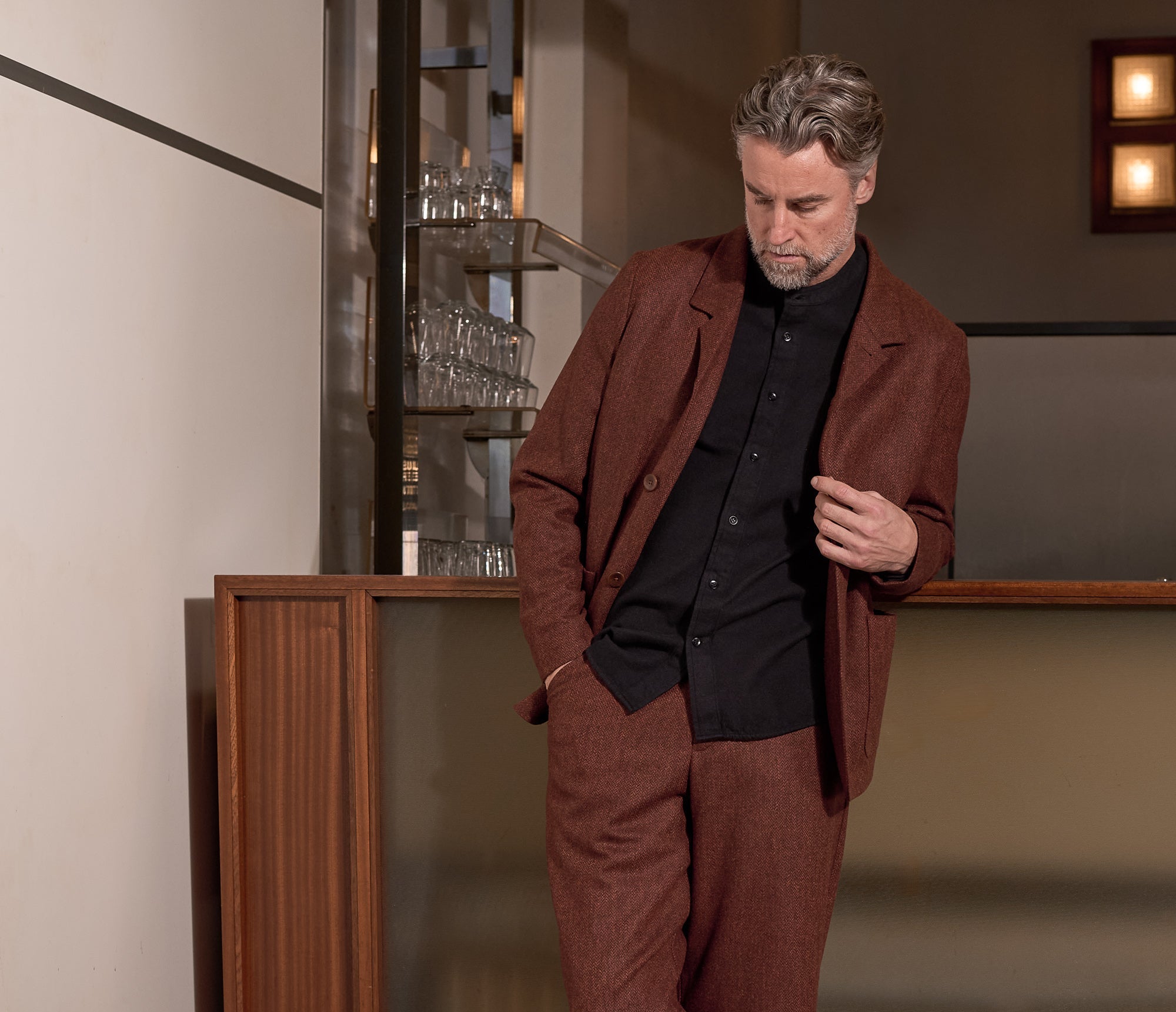 A man wearing a matching herrinbone brick lambswool blazer and trousers set with a black shirt and black shoes, leaning against a wooden counter in an indoor setting.