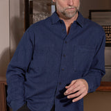 A man wearing a dark blue long-sleeve button-down shirt with a chest pocket, paired with dark corduroy trousers, standing in an indoor setting.